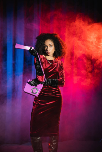 Fashionable african american model in velvet dress holding telephone on blue background with red light and smoke - Φωτογραφία, εικόνα