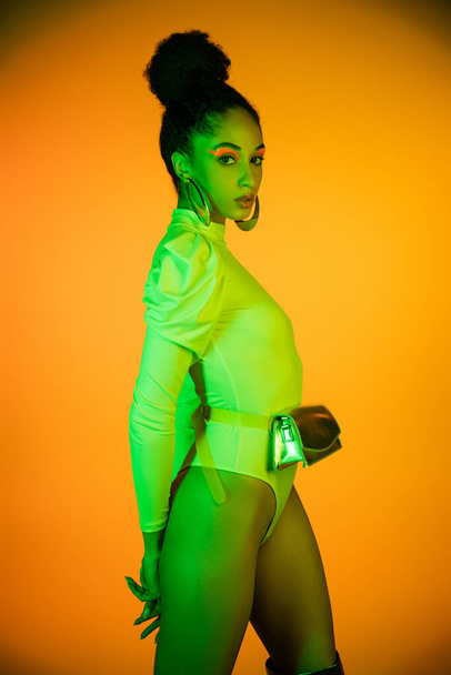 Stylish african american model with neon visage and bodysuit standing on orange background  - Фото, изображение