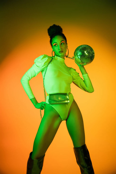 Trendy african american woman in neon bodysuit holding disco ball on orange background  - Photo, Image