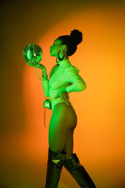 Seitenansicht der sexy afrikanisch-amerikanischen Frau im Neon-Body mit Blick auf die Discokugel auf orangefarbenem Hintergrund  - Foto, Bild