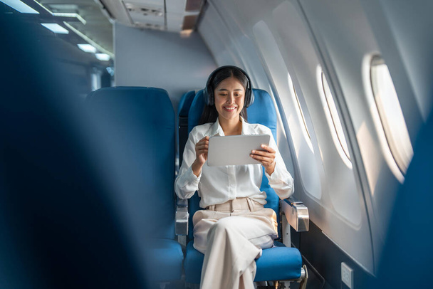 Νεαρή Ασιάτισσα επιχειρηματίας ή γυναίκα επιβάτης που φοράει ασύρματα ακουστικά και εργάζεται με tablet κατά τη διάρκεια της πτήσης. - Φωτογραφία, εικόνα