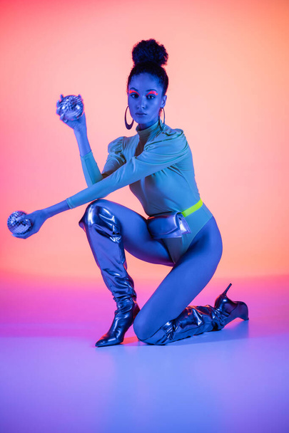 Trendy african american woman in bodysuit holding disco balls on pink and purple background with neon light  - Фото, зображення