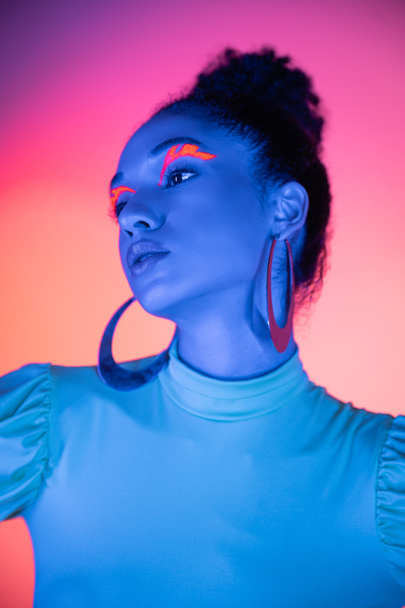 Young african american model with neon makeup looking away on pink and purple background - Foto, afbeelding