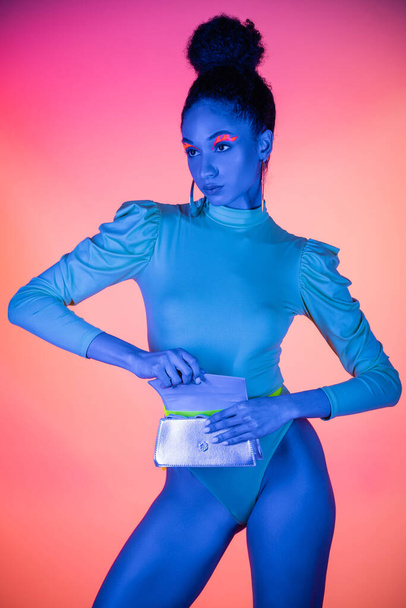 Pretty african american model with neon visage and bodysuit opening waist bag on light pink background - 写真・画像