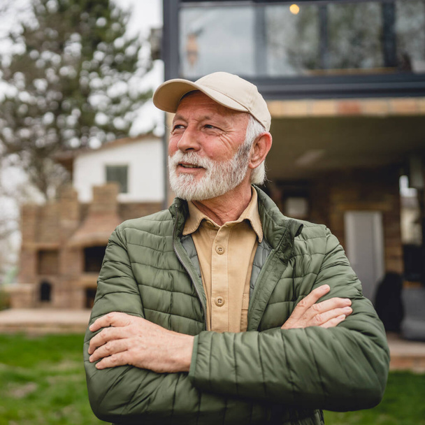 One modern senior caucasian man portrait of male grandfather with beard and hat cap stand in front of his house in day wear jacket happy confident copy space arms crossed - Photo, Image