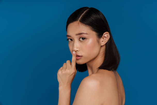 Portrait of young beautiful asian woman showing silence gesture with finger on lips and looking at camera, while standing with her back over isolated blue background - Fotoğraf, Görsel