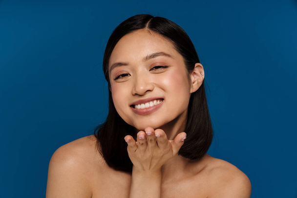 Young beautiful smiling happy asian woman showing air kiss gesture and looking at camera while standing over isolated blue background - Photo, image