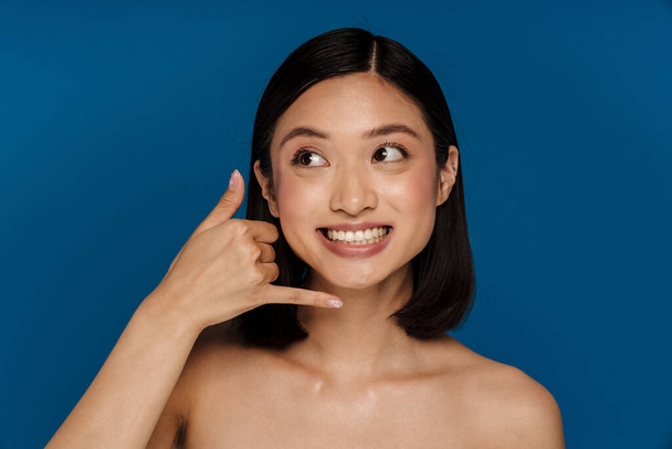 Young beautiful happy smiling asian woman doing phone gesture and looking aside, while standing over isolated blue background - Foto, imagen