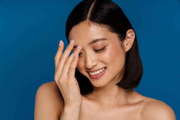 Junge schöne lächelnde glückliche asiatische Frau mit geschlossenen Augen, die ihren Nasenrücken berühren, während sie vor isoliertem blauen Hintergrund steht - Foto, Bild