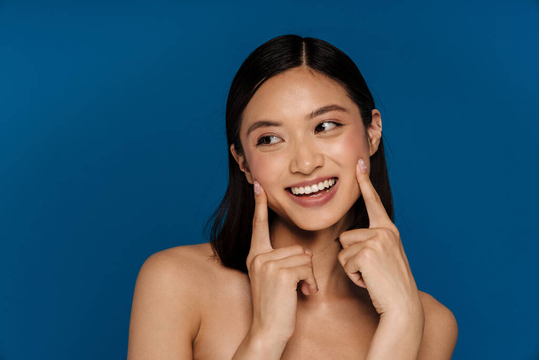 Young beautiful smiling happy asian woman touching her cheeks and looking atside , while standing over isolated blue background - Foto, Bild