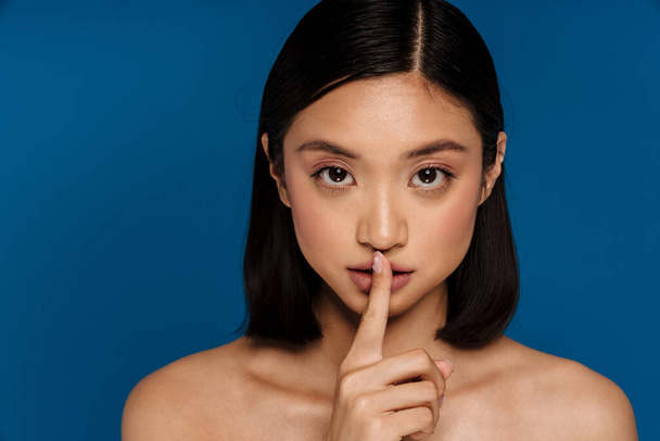 Young beautiful asian woman showing silence gesture with finger on lips and looking at camera, while standing over isolated blue background - Fotoğraf, Görsel