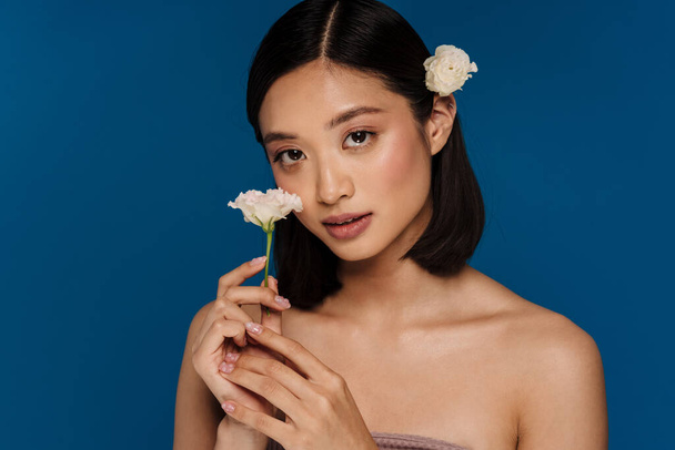 Portrait of young beautiful asian woman with opened mouth with flower on ear holding white flower and looking at camera, while standing over isolated blue background - Foto, Bild