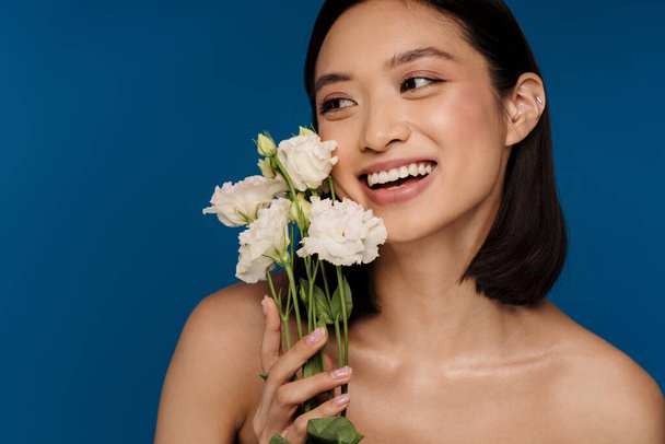 Portrait of young beautiful smiling happy asian woman with white flowers looking aside, while standing over isolated blue background - Foto, Bild