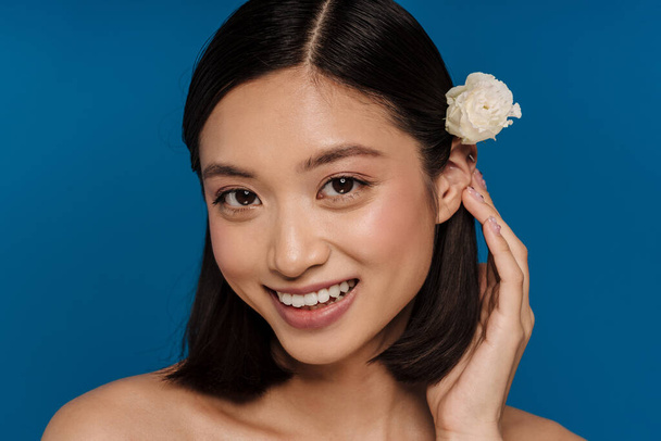 Portrait of young beautiful smiling asian woman with flower on ear looking at camera and adjusting her hair , while standing over isolated blue background - Foto, afbeelding
