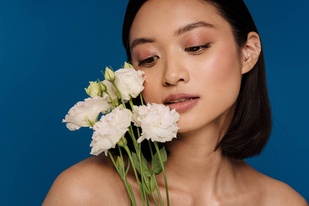 Portrait of young beautiful calm asian woman with white flowers looking downward, while standing over isolated blue background - Foto, Bild