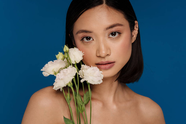 Portrait of young beautiful calm asian woman with white flowers looking at camera, while standing over isolated blue background - Φωτογραφία, εικόνα