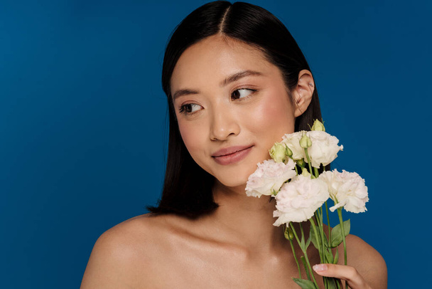 Portrait of young beautiful calm asian woman with white flowers looking aside, while standing over isolated blue background - Foto, afbeelding