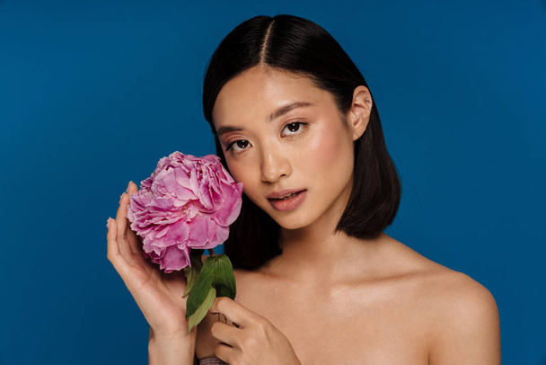 Asian half-naked woman looking aside while posing with peony isolated over blue background - Fotó, kép