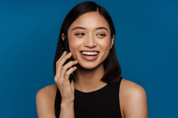 Asian young woman laughing while talking on mobile phone isolated over blue background - 写真・画像