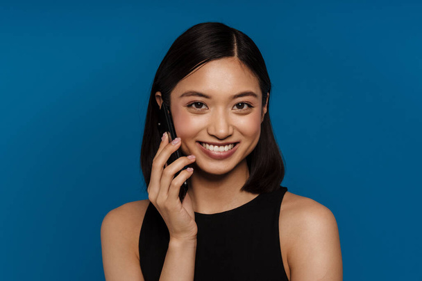 Asian young woman laughing while talking on mobile phone isolated over blue background - Fotoğraf, Görsel