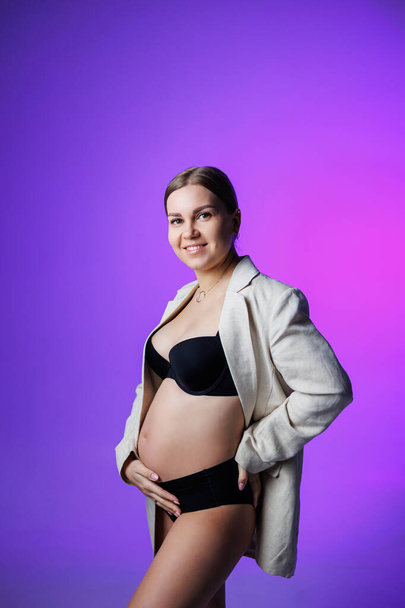 Ohut raskaana oleva nainen, jolla on pitkä takki ja alusvaatteet, seisoo studiossa valkoisella taustalla pitäen kätensä vatsallaan. Naisten alusvaatteet raskaana oleville naisille - Valokuva, kuva
