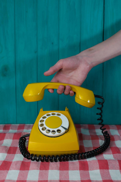 teléfono retro amarillo con un disco blanco y números negros y una mano con un receptor sobre un fondo de madera turquesa y sobre un tejido rojo de cuadros blancos - Foto, imagen