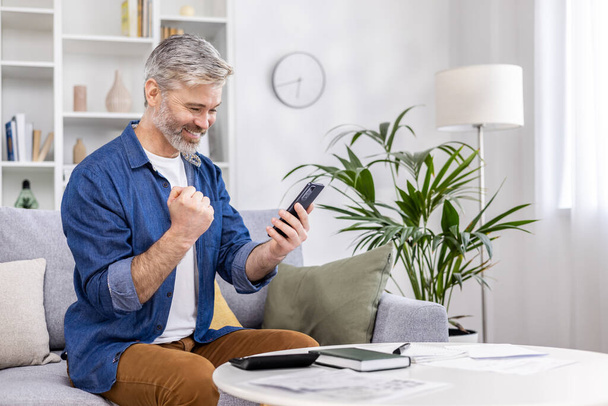 Senior gray-haired male businessman sitting on the sofa at home and using the phone. Received good news, financial success. Shows yes with his hand, celebrates, rejoices. - Fotó, kép