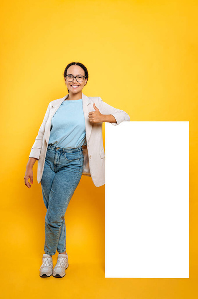 Full length vertical photo of a positive brazilian woman, stands near an square white mock-up poster for presentation, announcement or advertising, shows thumb-up, smiling, isolated yellow background - Valokuva, kuva