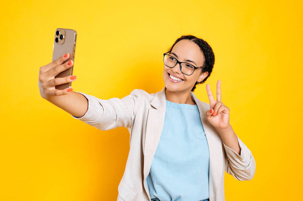 Joyful lovely pretty brazilian woman with glasses, stylishly dressed, holds smart phone in hand, looks at phone camera, takes a selfie photo, smile and show peace gesture, isolated yellow background - Fotografie, Obrázek