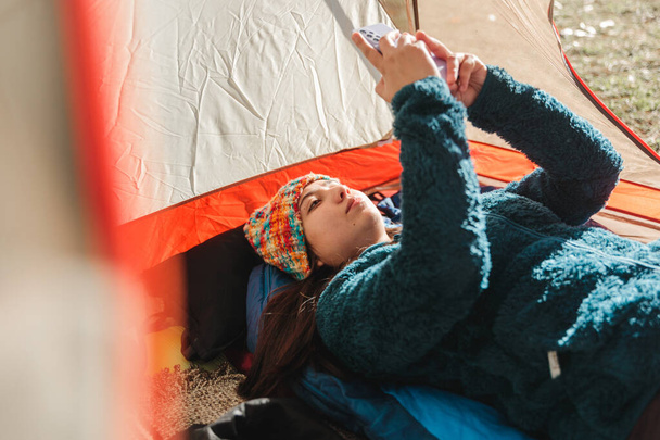 Junge Frau in warmer Kleidung, mit ihrem Handy, das morgens in einem Zeltlager liegt. Hochwertiges Foto - Foto, Bild