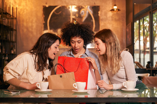 Kolme monirotuista ystävää kahvilassa tekemässä vuosipäivälahjoja. Säästökaupasta tulevat naiset katsovat pussia yllättyneenä kahvin äärellä. Innostuneet ihmiset saavat odottamattomia lahjoja. - Valokuva, kuva