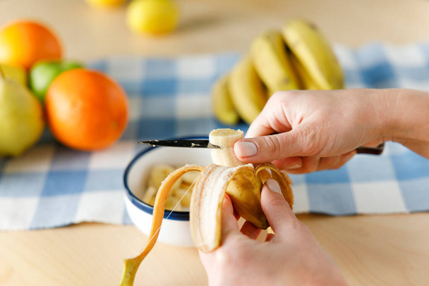 Woman sitting by the table at home cutting banana. She is preparing fruit salad at home  - Foto, Imagem