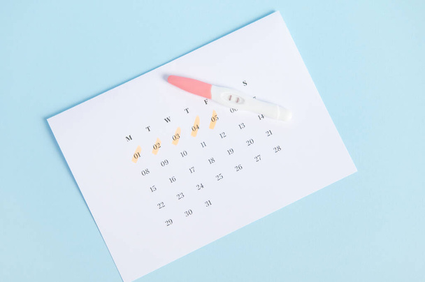Szybka diagnoza ciąży w domu. Widok z góry testu ciążowego atramentem z dwoma paskami w białym kalendarzu z zaznaczonymi datami ostatniej miesiączki, izolowane na niebieskim tle. Koncepcja zdrowia kobiet - Zdjęcie, obraz