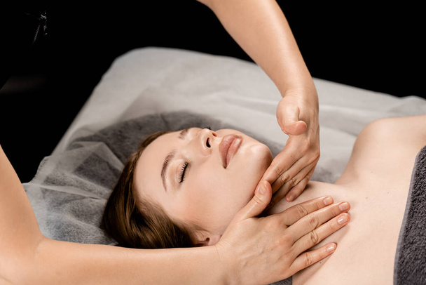 Girl with closed eyes is relaxing on face and neck massage in spa. Masseur is making facial massage for young woman in spa. Facebuilding and relaxing procedures - Photo, Image