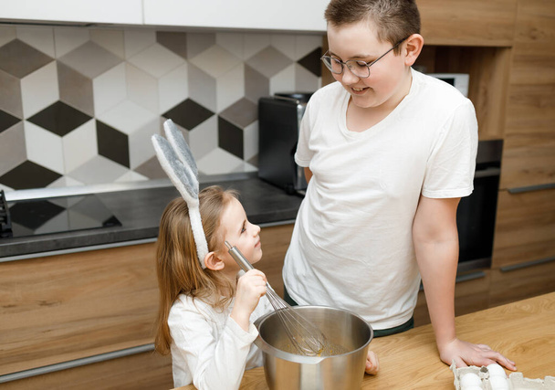 Αδελφός με γυαλιά και η μικρή αδελφή του στην κουζίνα χαμογελώντας και κοιτάζοντας ο ένας τον άλλον ενώ μαγειρεύουν. Παιδί ανακατεύει ζύμη σε αυτιά κουνελιού. Βοήθεια, έννοια της οικογενειακής υποστήριξης - Φωτογραφία, εικόνα