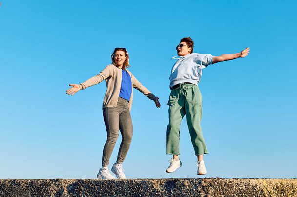 Две юные девушки танцуют, веселятся и дурачатся на большом камне на берегу моря с голубым небом. - Фото, изображение