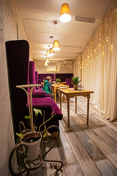 niño en un chaleco verde come un pastel en un interior moderno y elegante con sillas púrpuras y mesas de madera - Foto, Imagen