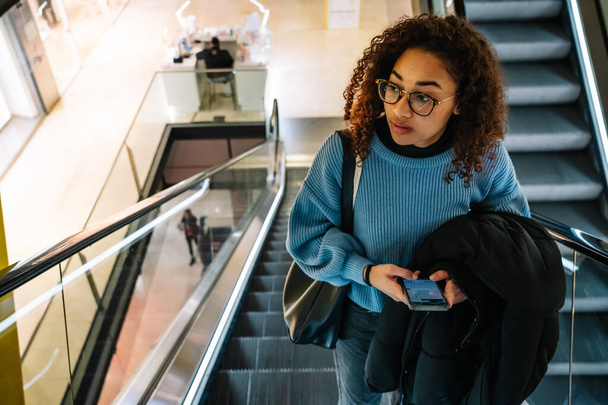 D'en haut de la jeune femme afro-américaine avec des cheveux bouclés dans des vêtements décontractés et des lunettes debout sur l'escalator et la navigation téléphone mobile dans le centre commercial - Photo, image