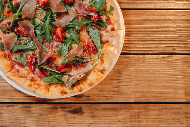 Auf dem Holztisch liegt ein Teller mit einer kleinen Pizza in der Pizzeria. Wir sehen viel Fleisch, Tomaten, Paprika, Käse. Es sieht erstaunlich lecker und schmackhaft aus. Es wird dem Kunden serviert. - Foto, Bild