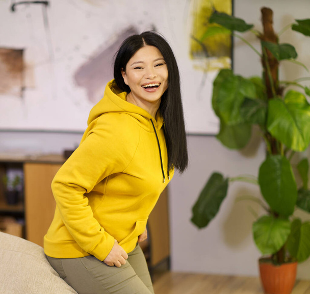 Happy Asian woman is leaning on the sofa and smiling at home. The image represents the concept of a happy and healthy lifestyle with a focus on biohacking. High quality photo - Zdjęcie, obraz