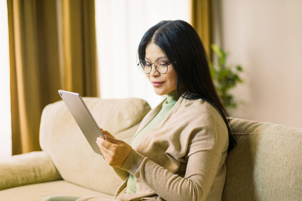 Mature Asian woman wearing glasses is deeply focused on browsing internet on her tablet while comfortably seated in the interior of her home. High quality photo - Foto, afbeelding