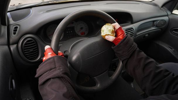mani sul volante di un'auto in guanti rossi, con una mela in una mano una persona guida un'auto. Concetto: Mangiare                       - Foto, immagini
