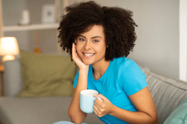 Mosolygó fiatal göndör afro-amerikai nő csésze forró ital ül a kanapén, élvezi a pihenést egyedül a nappali belsejében. Kávészünet, tea és pihenés otthon, szabadidőben - Fotó, kép
