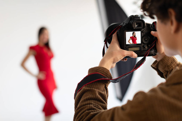 Profesyonel erkek fotoğrafçı ve kırmızı elbiseli güzel bir kadın model beyaz arkaplanda fotoşoplar yapıyor, kameraya odaklan. - Fotoğraf, Görsel