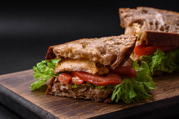 Delicioso sándwich con tostadas crujientes, pollo, tomates y lechuga sobre un fondo de hormigón oscuro - Foto, Imagen