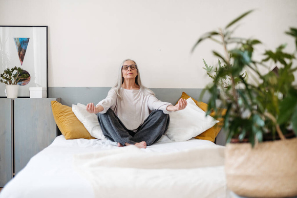 Senior woman doing yoga and meditating on a bed. - Фото, изображение