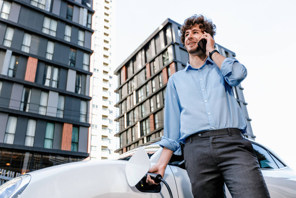 Progressive businessman talking on the phone, leaning on electric car recharging with public EV charging station, apartment condo residential building on the background as green city lifestyle. - Foto, imagen