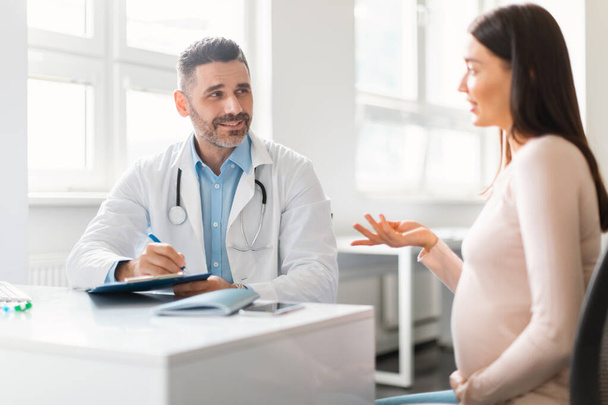 Nuori raskaana oleva nainen istuu sairaalassa, puhuu ja ottaa yhteyttä keski-ikäinen mies gynekologi. Terveydenhuollon ja lääketieteen käsite - Valokuva, kuva