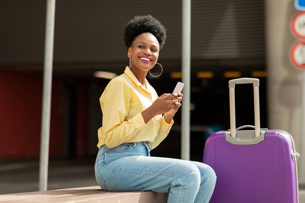 Podekscytowana czarna kobieta Turysta Korzystanie ze smartfona Przeglądanie Internetu i rezerwacji Bilety w aplikacji mobilnej Siedząc z walizką podróżną na zewnątrz w pobliżu lotniska. Turystyka i komunikacja - Zdjęcie, obraz