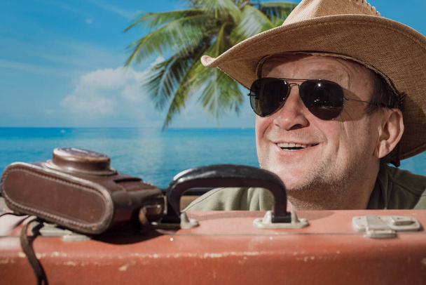 Happy aged man in sunglasses and hat with suitcase and photo camera on tropical beach next to sea, palm tree and blue sky background on sunny summer day in tropical beach. Vacation and travel concept - Φωτογραφία, εικόνα